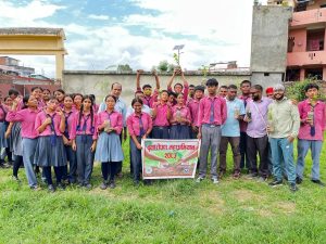 Students in pink uniforms and teachers holding a tree plantation campaign banner and saplings in a school ground