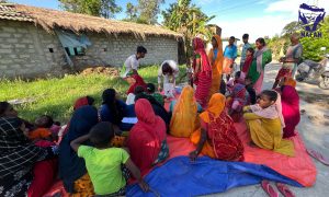 A woman handing seed to women gathered in colourful saris outdoors under blue sky