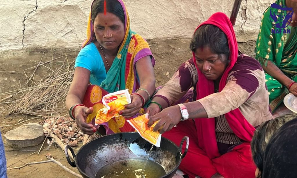 Women pouring spices into a large karahi of oil beside a pile of firewood